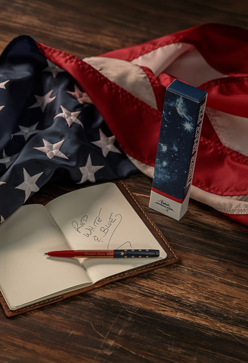 Patriotic product lifestyle scene featuring the Fisher Space Pen M4250.  A red and navy blue pen rests on an open leather notebook with 'Red White & Blue' handwritten on the page. Next to it stands the gift box with a cosmic space design. An American flag