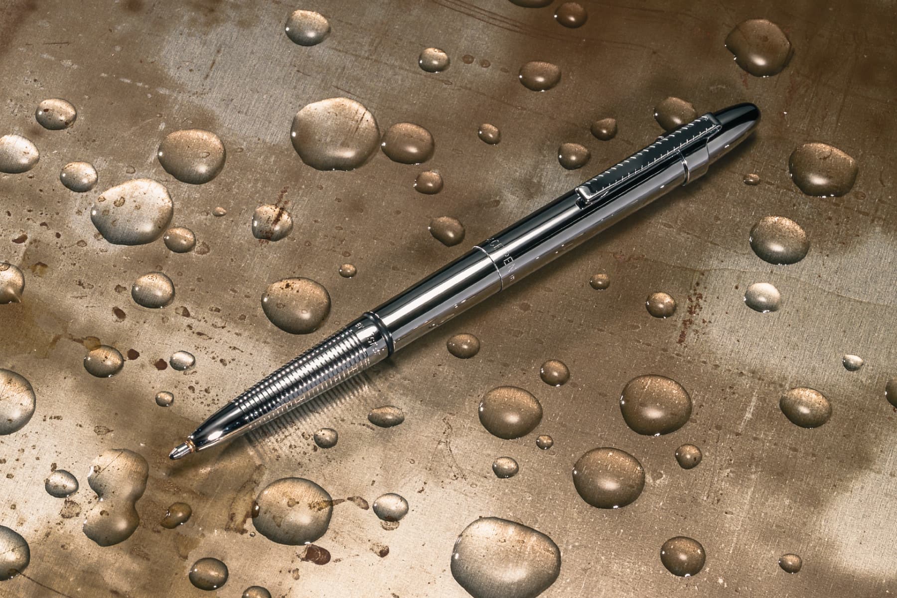 Open 400CL chrome bullet space pen with chrome clip laying atop a metal surface with water beading on it.