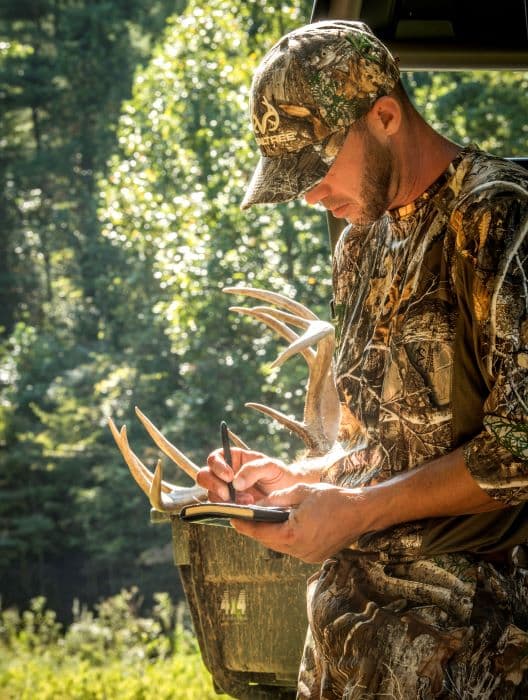 A man in camouflage clothing writing in a notebook with a 400H-237 Tungsten Cerakote bullet space pen. Behind him is a truck bed, antlers, and sunny trees and grass.