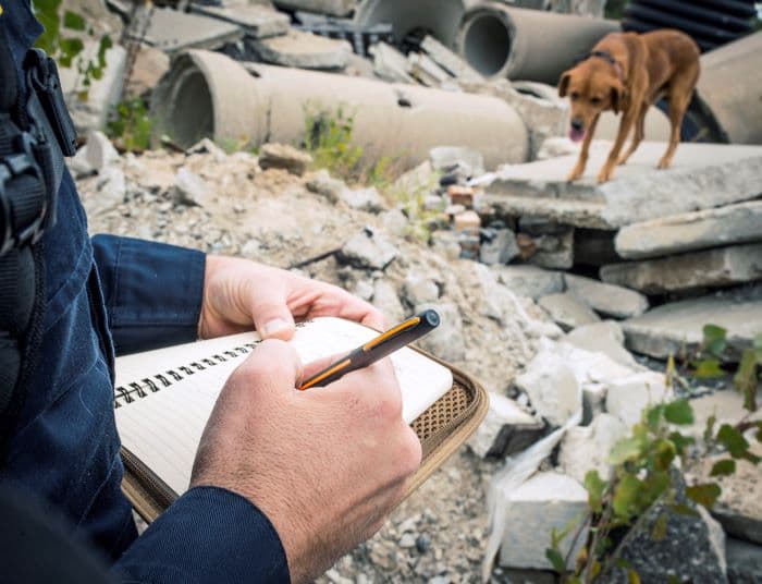 A person is writing in a lined notebook using the thin orange line matte black cap-o-matic Space Pen. In the background is a brown search dog standing atop concrete rubble.